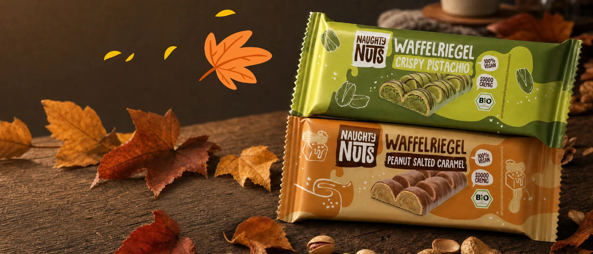 Zwei Naughty Nuts Waffelriegel in den Sorten Crispy Pistachio und Peanut Salted Caramel auf einem Holztisch mit Laub
